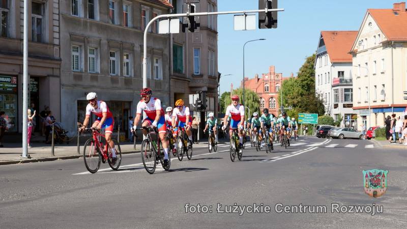 Na pierwszym planie grupa rowerzystów. Na początku grupy kilku w biało-czerwono-niebieskich strojach, a z tyłu w zielono-niebieskich. W tle widać budynki miejskie oraz sygnalizacje świetlne.